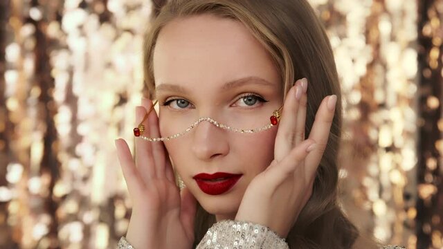 Fashion luxury portrait of blonde model emotionally looking at camera with red lips makeup, sparkling jewelry earrings and crystal glasses on festive glittering background. 
