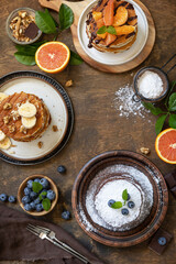 Celebrating Pancake day, healthy breakfast. Delicious homemade american bananas pancakes with blueberries, nuts, caramel, chocolate, red orange on rustic wooden table. Top view flat lay.