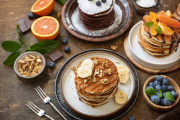 Celebrating Pancake day, healthy breakfast. Delicious homemade american bananas pancakes with blueberries, nuts, caramel, chocolate, red orange on rustic wooden table.