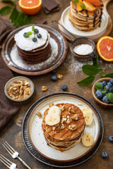 Celebrating Pancake day, healthy breakfast. Delicious homemade american bananas pancakes with blueberries, nuts, caramel, chocolate, red orange on rustic wooden table.