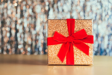 Glittering golden gift box with velvet red ribbon with a bow on the sparkling gold tinsel background. The concept of a gift for birthday, Valentine's day, Xmas, new year. Festive background.
