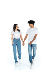 full length view of happy interracial couple holding hands and looking at each other while walking on white.