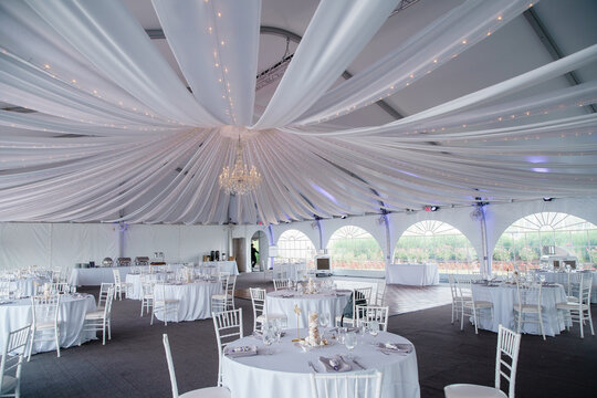 A wide-angle view of a wedding reception venue with dance floor and surrounding tables A large chandelier in the middle of the room Tooling draped from it with mini lights white lacy fabric spread