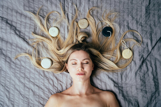 View from above of blond woman close eyes lying on bed with natural eco friendly solid shampoo bars or conditioners on her hair. Zero waste and sustainable lifestyle.