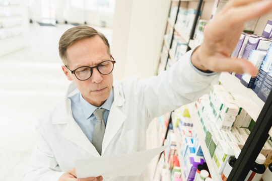 Male Caucasian Middle-aged Mature Druggist Pharmacist Looking Reading Prescription For Checking Expiration Date, Side Effects, Active Substance Of Drugs, Pills, Medicines In Pharmacy