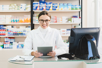 Fototapeta na wymiar Smiling cheerful caucasian female young pharmacist druggist holding tablet for checking drugs medicines pills remedies on at cash point desk in drugstore pharmacy
