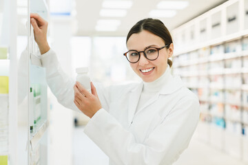 Caucasian female young pharmacist druggist in white medical coat holding remedy pills jar while looking at camera in pharmacy drugstore