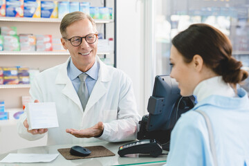 White blank medicine box container with drugs, vitamins, pills, painkillers, antibiotics. Middle-aged mature male pharmacist showing advising medications to female client customer