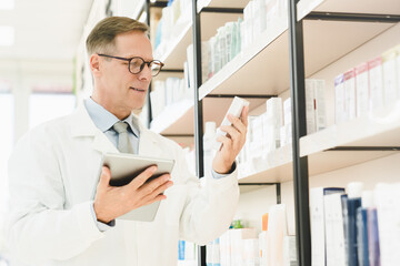 Naklejka na ściany i meble Male caucasian middle-aged mature pharmacist druggist holding medicines jar of pills drugs remedy checking its ingredients using digital tablet in drugstore pharmacy