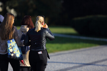 Dwie kobiety, dziewczyny spacerują po chodniku w parku, piją kawę z kubka, telefon w kieszeni.