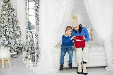 A happy mother is having fun with her son, who opens a wrapped present at home during the celebration. Mother and son are sitting on a white bed and opening a red gift box with a Christmas present.