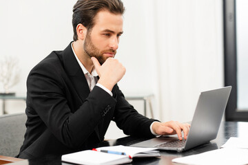 Handsome focused young businessman in formal wear using laptop computer sitting at the desk in office, charismatic bearded male employee checking emails, man in suit watching at screen thoughtfully