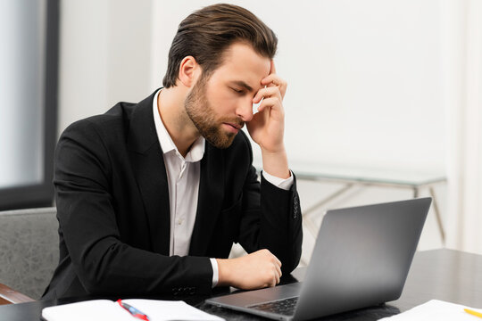 Frustrated young businessman in formal suit made mistake and worried about it, tired male office employee suffering from strong headache, sitting at the desk in office with eyes closed