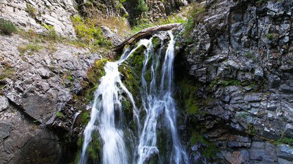 A mountain waterfall in a rocky gorge. The view from the drone is of steep cliffs, moss, green plants and the river. There are stones lying. Splashes from the water. There is a log lying. Almaty
