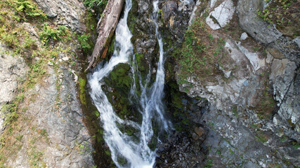 A mountain waterfall in a rocky gorge. The view from the drone is of steep cliffs, moss, green plants and the river. There are stones lying. Splashes from the water. There is a log lying. Almaty