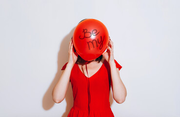 Valentine's Day. Portrait of a beautiful young woman in red dress holding red balloon on white background. Be my Valentine