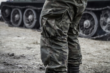 Fototapeta na wymiar Soldier's legs on a battlefield, wearing woodland camo military pants (camouflage trousers). With a tank tracked wheels in the background.