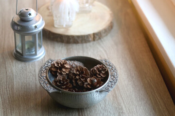 Silver bowl filled with natural pinecones, lantern and lit candles on the table. Hygge at home. Selective focus.