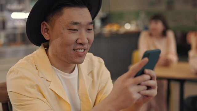 Portrait of smiling asian guy using smartphone at cafe