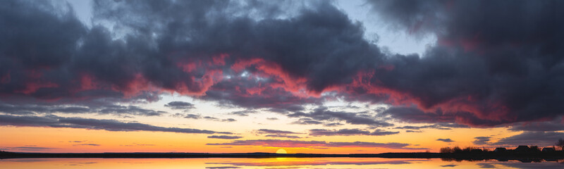 A bright sunset with a large yellow sun reflected in the water. Sunrise at the edge of the village. The big sun goes over the horizon reflecting the light in the lake