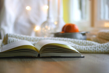 Open book, bowl of oranges, knitted blanket and lit candles on the table. Hygge at home. Selective focus.