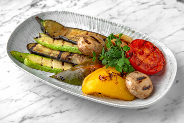 Grilled vegetables: zucchini, tomato, pepper and mushrooms in a festive plate on a marble background. Restaurant banquet menu.