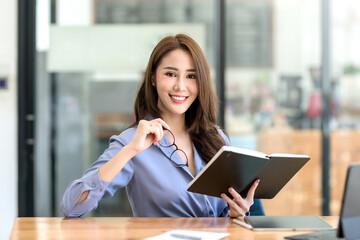 Beautiful young Asian businesswoman sitting holding a book looking at camera with laptop in office.