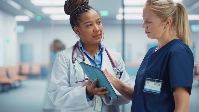 Medical Hospital: Professional Black Female Medical Doctor Talks with White Female Nurse Using Digital Tablet Computer. Two Health Care Specialists Discuss, Find Treatment Solution to Save Lives