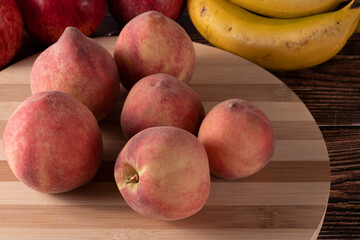 Tasty peaches (Prunus persica, in Latin), on wooden board, on rustic wooden table, with banana and apple in the background. Selective focus. Top view.