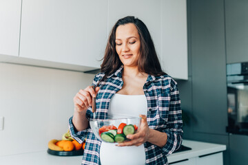 Portrait of young smiling pregnant woman mix vegetable salad with cucumbers and tomatoes in transparent bowl in kitchen.