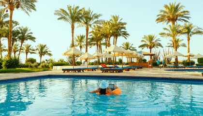 A woman in a swimsuit and a sun hat in the pool in a swimming circle with a laptop with her back to the camera on a background of palm trees.