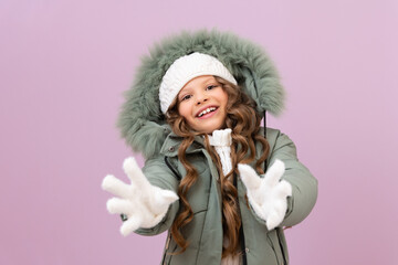 a little girl in winter clothes and curly hair pulls her hands forward and smiles. kid in a warm winter jacket and a white hat on a purple isolated background.