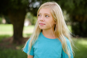 Caucasian little kid girl wearing blue T-shirt standing outdoors looking aside into empty space thoughtful