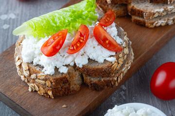 Slice of rye bread with cottage cheese and tomatoes on a wooden cutting board on a gray background