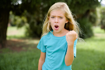 Caucasian little kid girl wearing blue T-shirt standing outdoors angry and mad raising fist frustrated and furious while shouting with anger. Rage and aggressive concept.