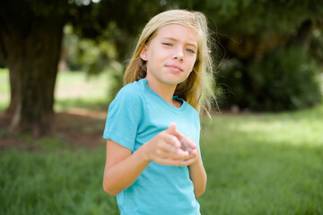 Joyful Caucasian little kid girl wearing blue T-shirt standing outdoors wink and points index...