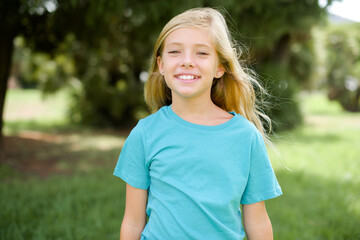 Caucasian little kid girl wearing blue T-shirt standing outdoors with a happy and cool smile on face. Lucky person.