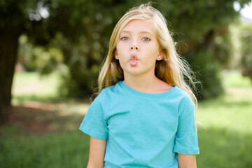 Caucasian little kid girl wearing blue T-shirt standing outdoors making fish face with lips, crazy and comical gesture. Funny expression.