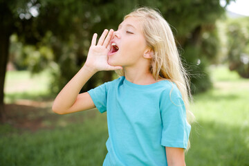Caucasian little kid girl wearing blue T-shirt standing outdoors shouting and screaming loud to side with hand on mouth. Communication concept. - obrazy, fototapety, plakaty