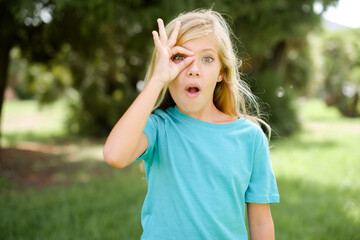 Caucasian little kid girl wearing blue T-shirt standing outdoors doing ok gesture shocked with surprised face, eye looking through fingers. Unbelieving expression.