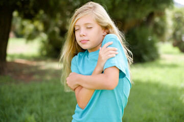 Caucasian little kid girl wearing blue T-shirt standing outdoors Hugging oneself happy and positive, smiling confident. Self love and self care