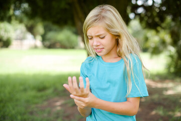 Caucasian little kid girl wearing blue T-shirt standing outdoors Suffering pain on hands and fingers, arthritis inflammation - obrazy, fototapety, plakaty