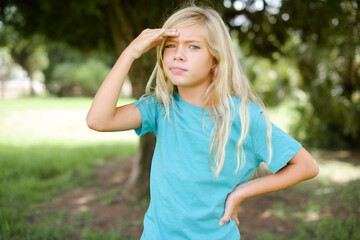 Caucasian little kid girl wearing blue T-shirt standing outdoors having problems, worried and stressed holds hand on forehead.