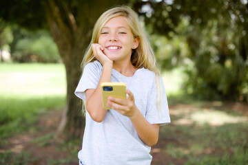 Joyous Caucasian little kid girl wearing white T-shirt poses with mobile phone device, types text message on modern smartphone, watches funny video during free time, enjoys good internet connection,