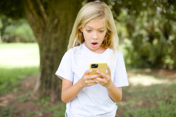 Focused Caucasian little kid girl wearing white T-shirt standing outdoors use smartphone reading social media news, or important e-mail