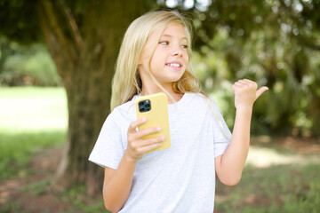 Caucasian little kid girl wearing white T-shirt standing outdoors using and texting with smartphone  pointing and showing with thumb up to the side with happy face smiling