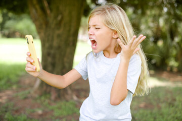 Angry Caucasian little kid girl wearing white T-shirt standing outdoors screaming on the phone,...