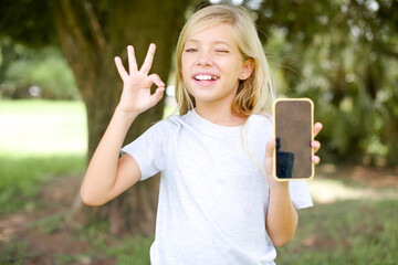 Caucasian little kid girl wearing white T-shirt standing outdoors holding in hands cell showing...