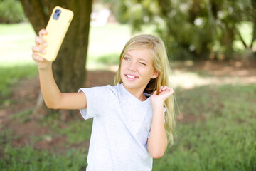 Caucasian little kid girl wearing white T-shirt standing outdoors smiling and taking a selfie ready...