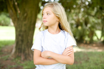 Charming thoughtful Caucasian little kid girl wearing whiteT-shirt standing outdoors stands with arms folded concentrated somewhere with pensive expression thinks what to do